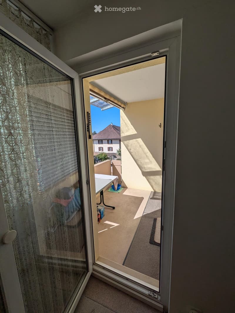 Open sliding glass door leads to a small balcony with a table and chair, overlooking a residential area with a visible house and clear blue sky.