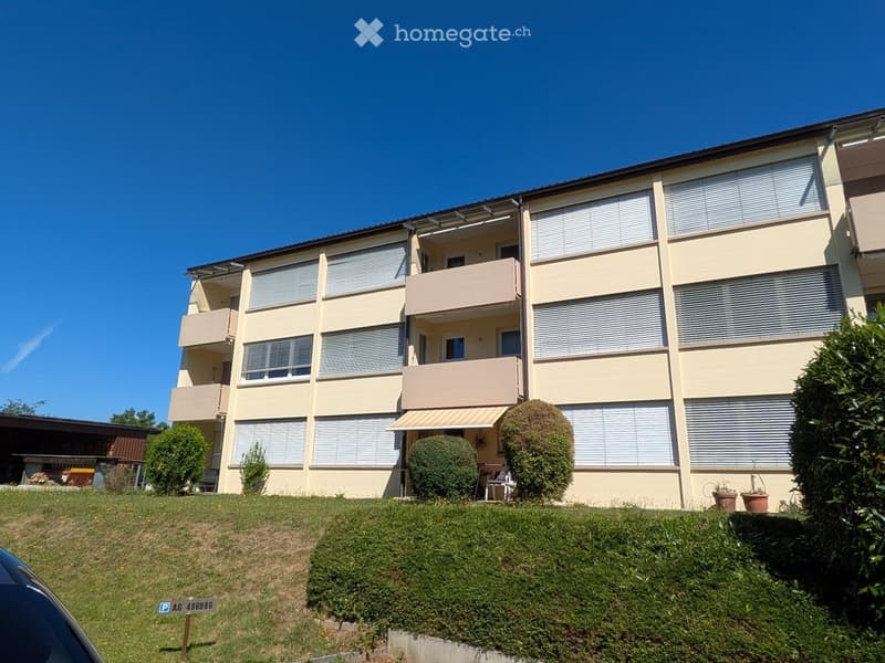 A multi-story residential building with beige walls, multiple windows, and balconies. The building is surrounded by greenery and has a well-maintained lawn in front.