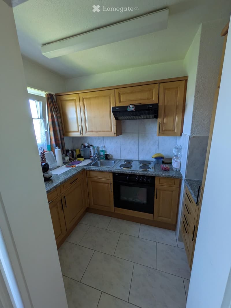 A compact kitchen with wooden cabinets, a tiled floor, a sink, stove, and various small appliances and items on the countertops.