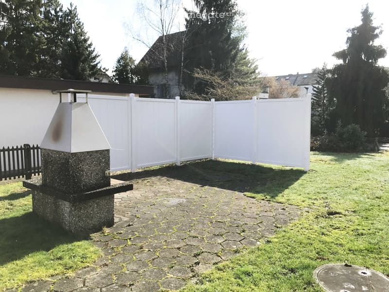 Garden area with a stone chimney, paved patio, white fence, and surrounding greenery.