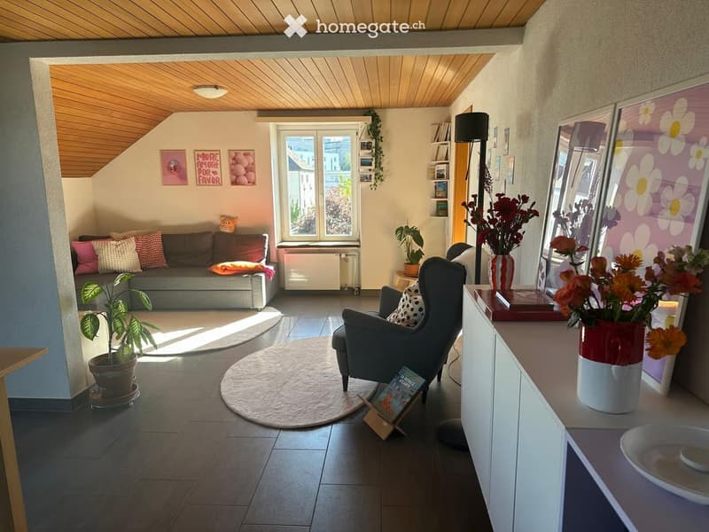 A cozy living room with a grey sofa, armchair, coffee table, and decorative vases. The room features wooden flooring, a large window with a view, and several potted plants.
