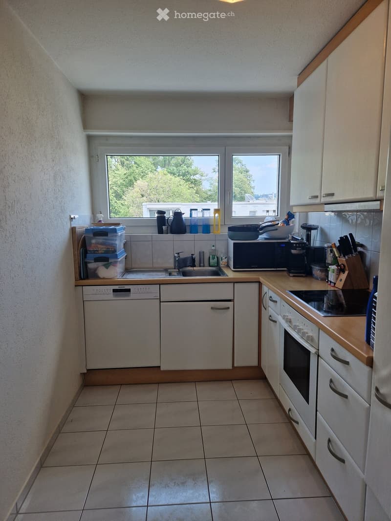 Modern kitchen with white cabinets, tiled floor, and various appliances including a microwave, coffee maker, oven, and dishwasher. The counter has a sink, cutting board, and various kitchen items.