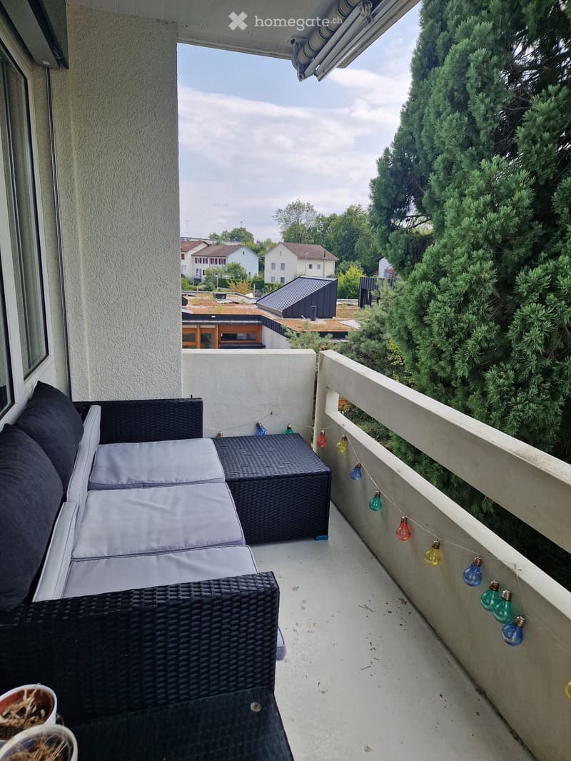 Balcony with a wicker sofa, small table, and potted plant, decorated with string lights and overlooking a residential area with trees.