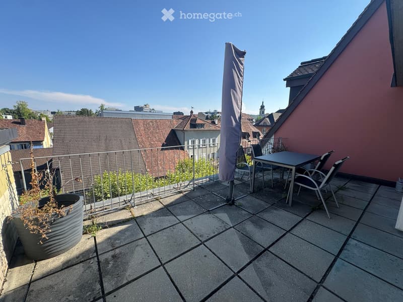 Balcony with tiled floor, metal railing, small table, chairs, and an umbrella. Potted plant visible on left side.