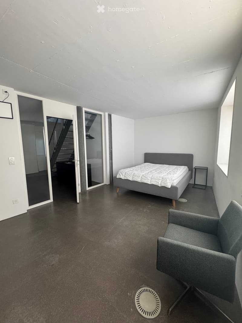 Modern bedroom with a grey bed, white bedding, grey chair, and a nightstand. The room features dark flooring and white walls, with a visible staircase in the background.