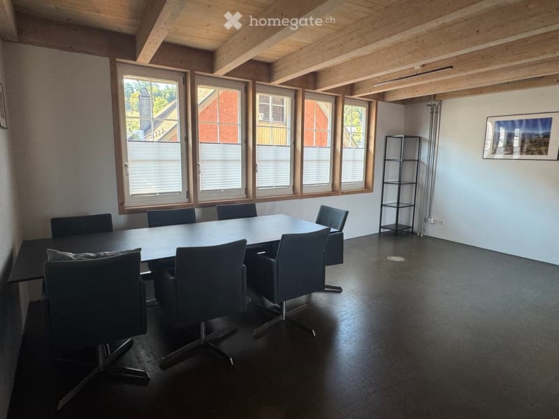 Bright, modern room with exposed wooden beams, large windows, a long conference table surrounded by chairs, and a tall, narrow shelving unit against the wall.