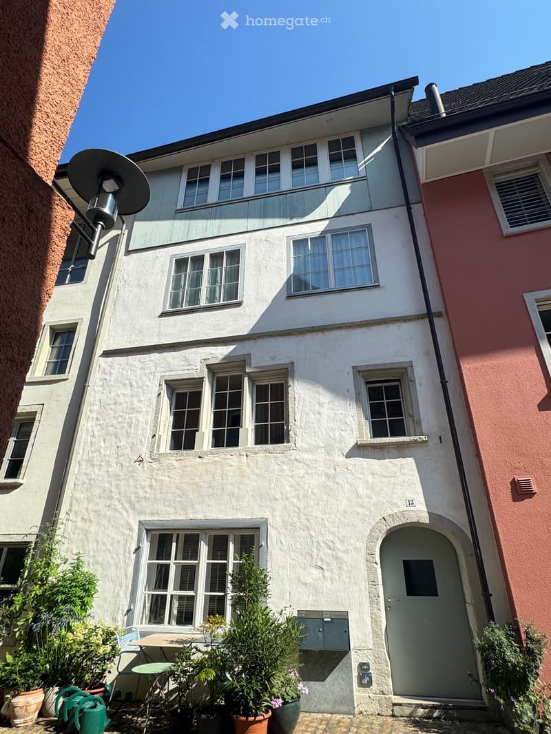 A three-story residential building with white plaster walls, multiple windows, and a small garden area with potted plants and a table in front of the entrance.