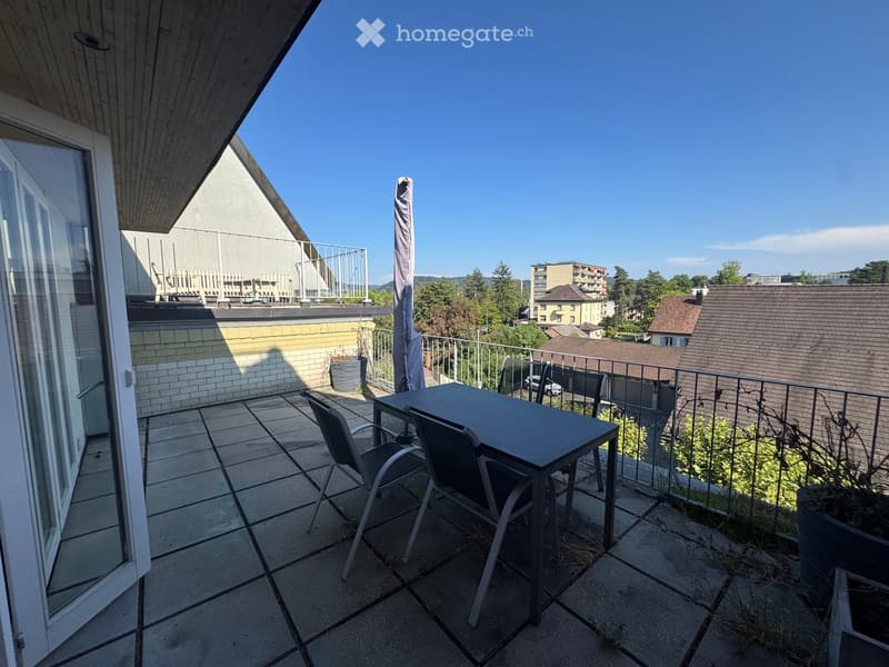 Balcony with tiled floor, metal railing, table, and chairs, overlooking a residential area with buildings and trees.