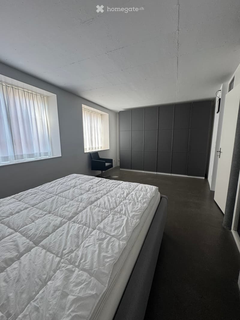 Modern bedroom with a large bed, a single chair, and a tall wardrobe. The room features grey walls, dark flooring, and two windows with white curtains.