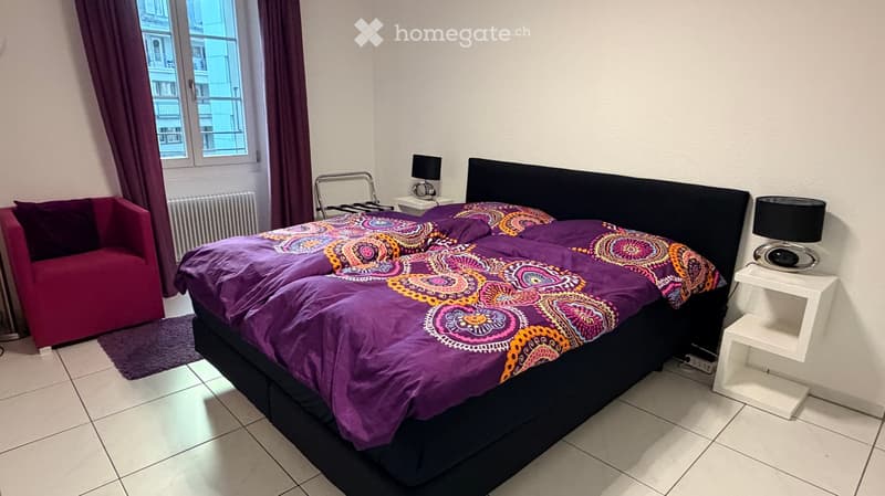 A modern bedroom with a large bed covered in a colorful, patterned duvet, a red armchair, and two nightstands with lamps.
