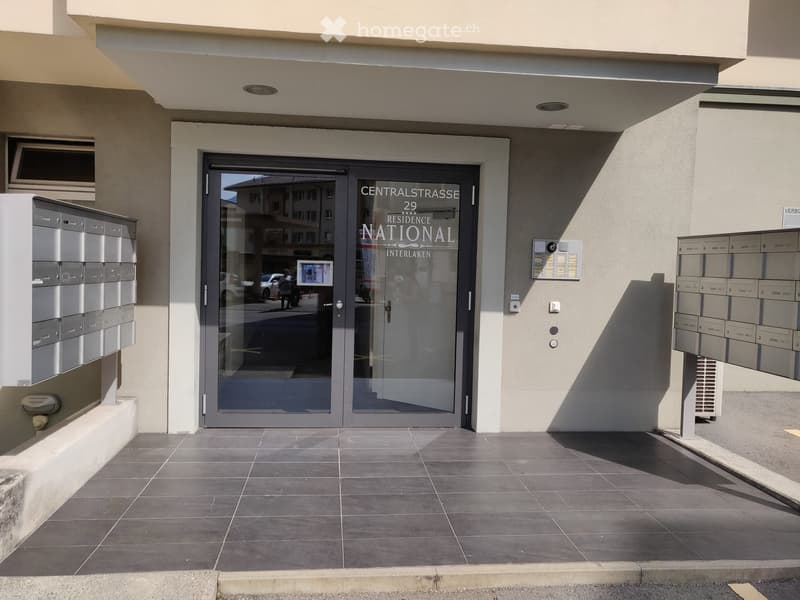 Entrance to a residential building with a glass door labeled "CENTRALSTRASSE 29 RESIDENCE NATIONAL INTERLAKEN" and mailboxes on either side.