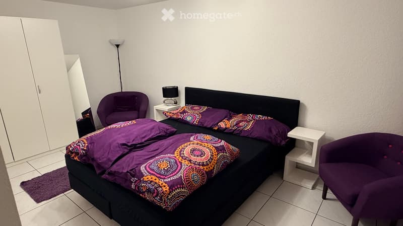 A modern bedroom with a double bed, purple bedding, two armchairs, a nightstand, and a large wardrobe.