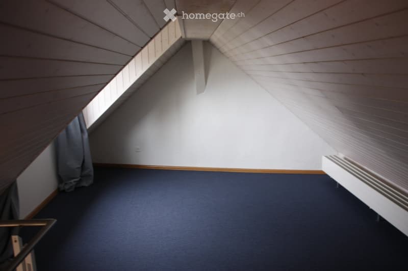 Empty attic room with wooden slanted ceilings, blue carpeted floor, white walls, and a small window with a curtain.