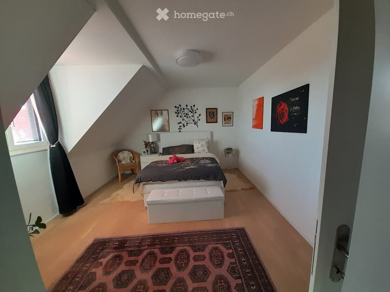 Cozy bedroom with slanted ceilings, wooden flooring, and a large bed with dark bedding. Includes a nightstand, chair, stool, and artwork on the walls.