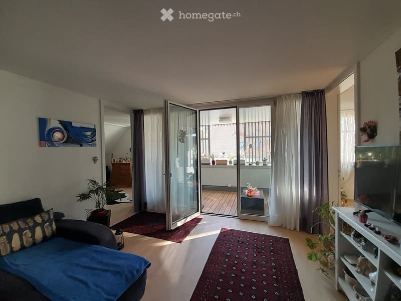 Bright living room with wooden flooring, large sliding glass doors leading to a balcony, a blue sofa, a coffee table, a television on a stand, and several plants.