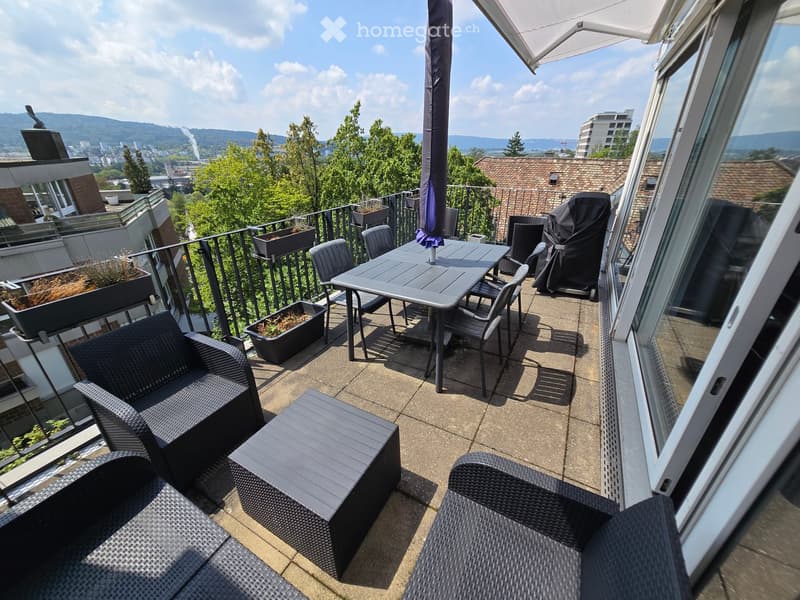 Balcony with outdoor furniture including a table, chairs, sofa, and umbrella, overlooking a cityscape with trees and buildings.