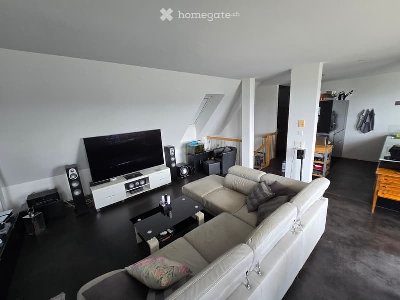 Modern living room with a white sectional sofa, coffee table, large flat-screen TV, speakers, and a fish tank. The room features dark flooring and white walls with a skylight window.