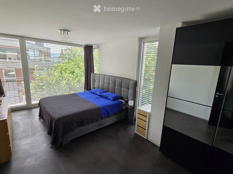 Modern bedroom with a large bed, nightstand, wardrobe, and large windows with blinds. The room features a tufted headboard and a sleek, contemporary design.