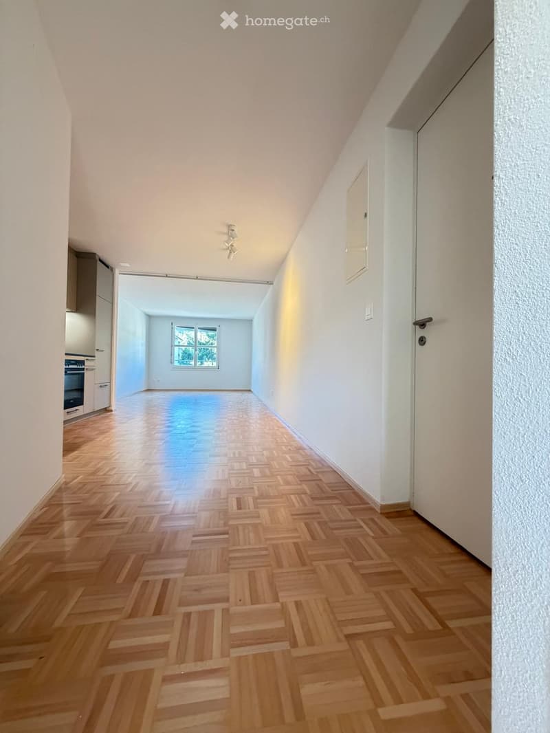 Empty hallway with wooden herringbone flooring, white walls, and recessed lighting. A door leads to a room with a window and a glimpse of a kitchen area with cabinets and an oven.