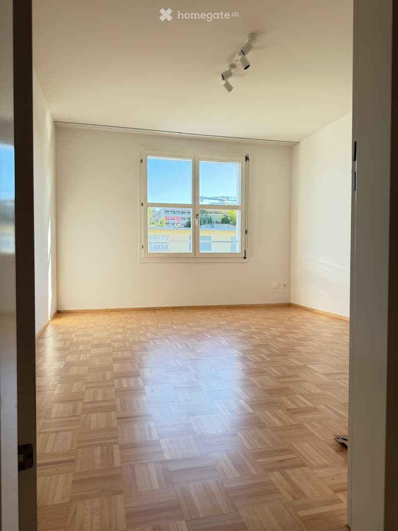Empty room with wooden flooring and white walls, featuring a large window with a view of the city and a track lighting fixture on the ceiling.