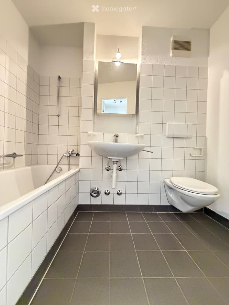 Modern bathroom with white tiled walls, dark grey tiled floor, bathtub, toilet, sink, and mirror.