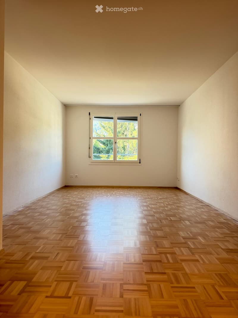 Empty room with wooden parquet flooring, white walls, and a large window with a view of greenery outside.