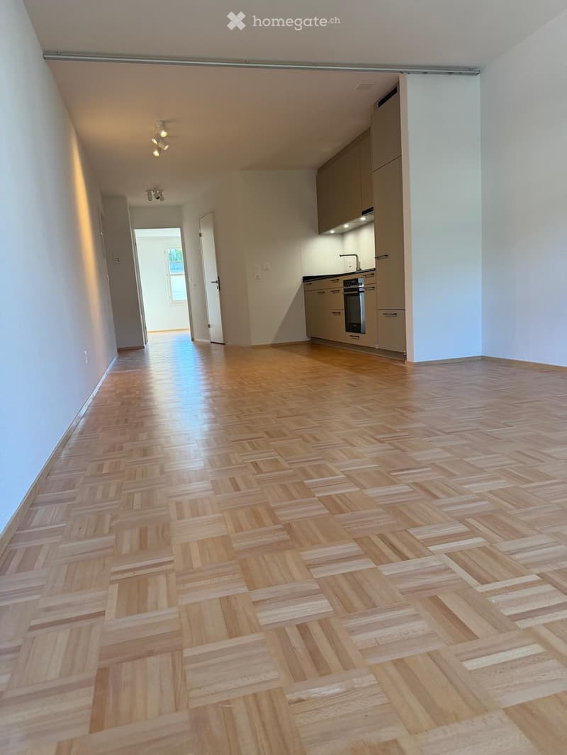 Empty hallway with wooden herringbone flooring, white walls, and recessed lighting. Kitchen area visible with cabinets and appliances.