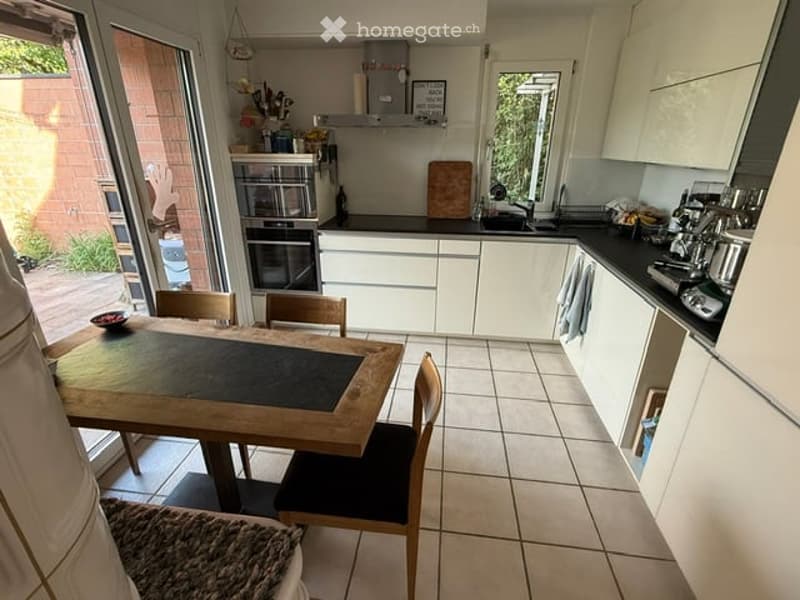 Bright, modern kitchen with white cabinets, a wooden dining table with chairs, an oven, a fridge, and a sink. The space features tiled flooring and ample natural light from large windows.
