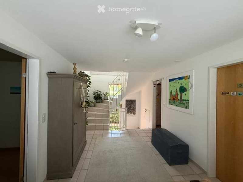 Bright hallway with white walls, tiled floor, and a staircase leading upwards. Features include a dark wooden bench, a grey cabinet, and a framed picture on the wall.