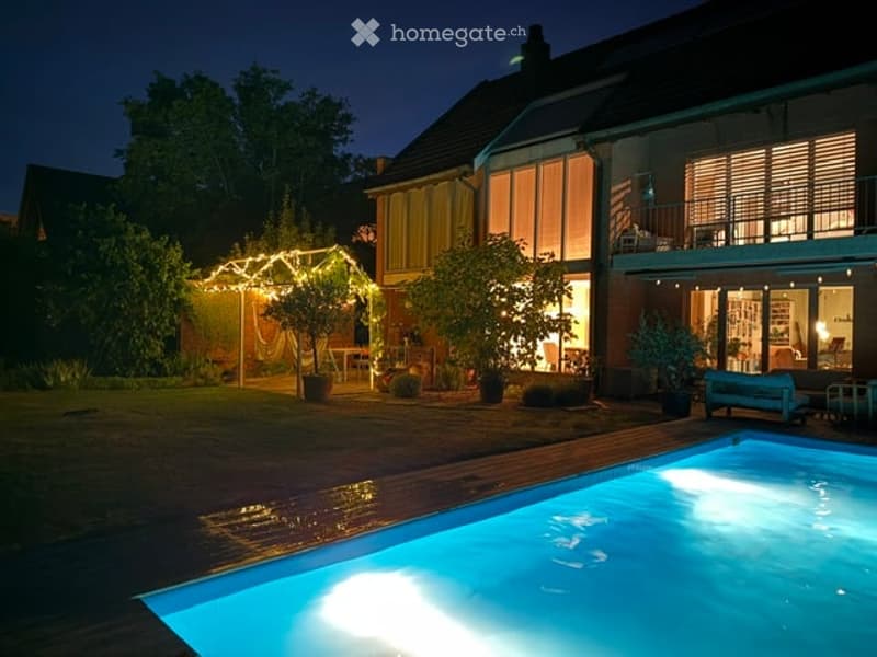 Nighttime view of a modern house with a swimming pool in the foreground, surrounded by a well-maintained garden with string lights and potted plants.