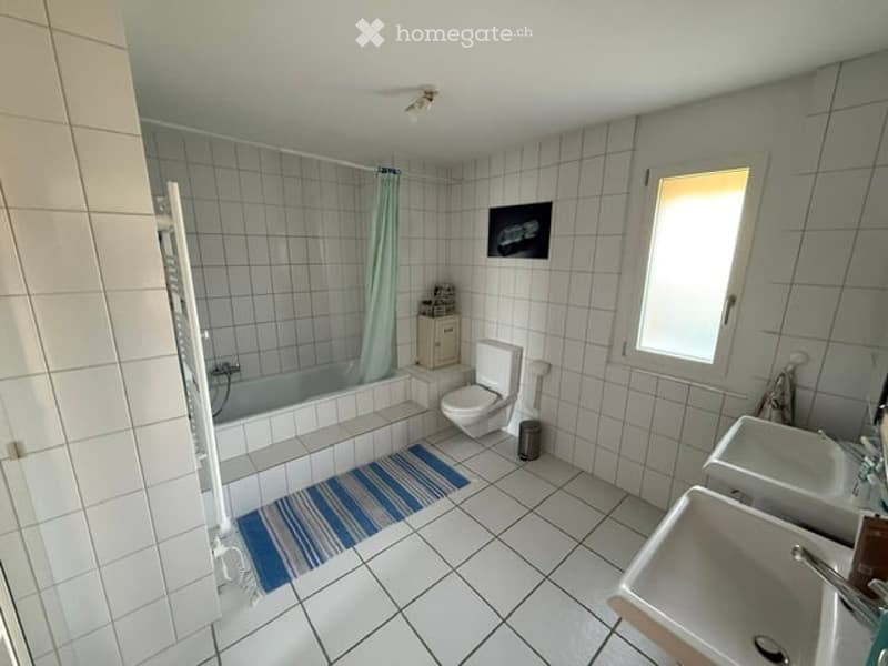 Bright bathroom with white tiled walls and floor, featuring a bathtub, toilet, double sink, and a small cabinet. A window provides natural light, and a striped rug is placed in front of the bathtub.