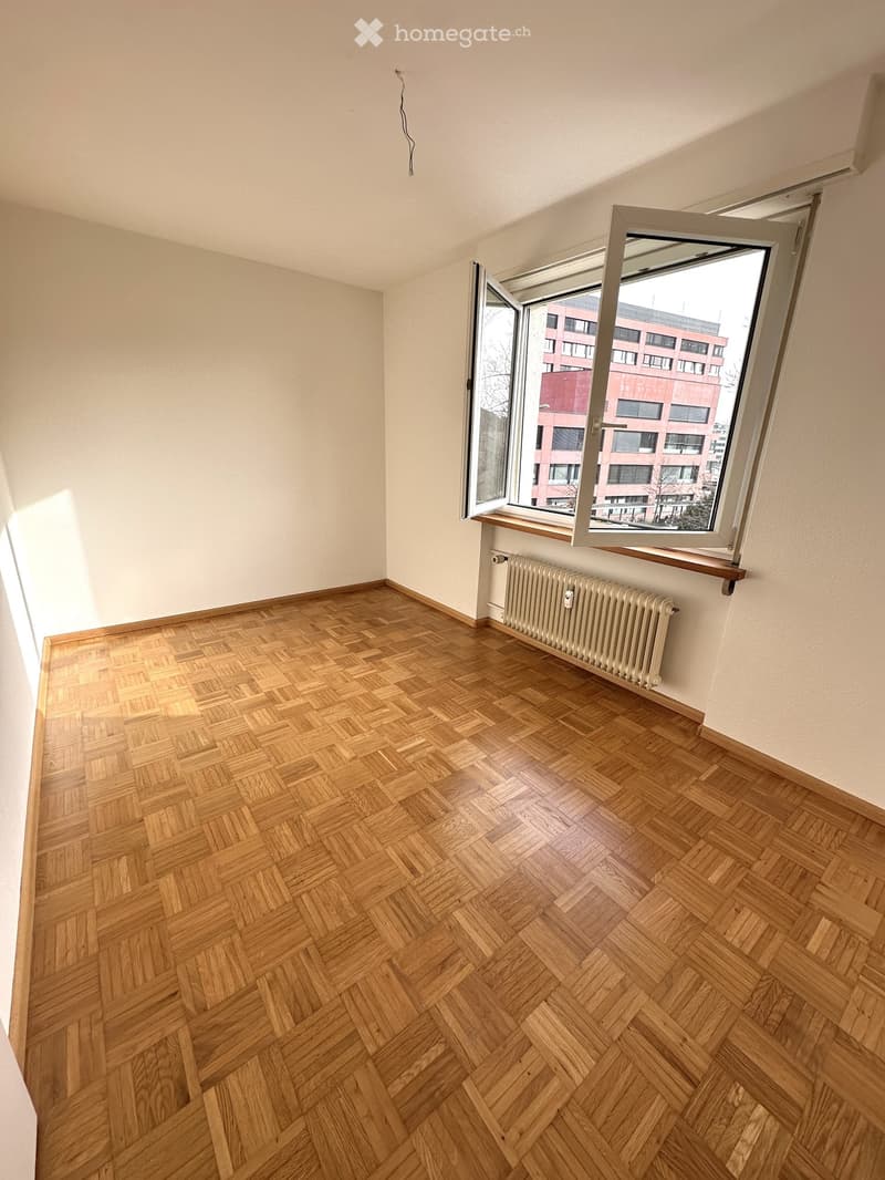 empty room, wooden parquet flooring, large window with a view of a red building, white walls, radiator