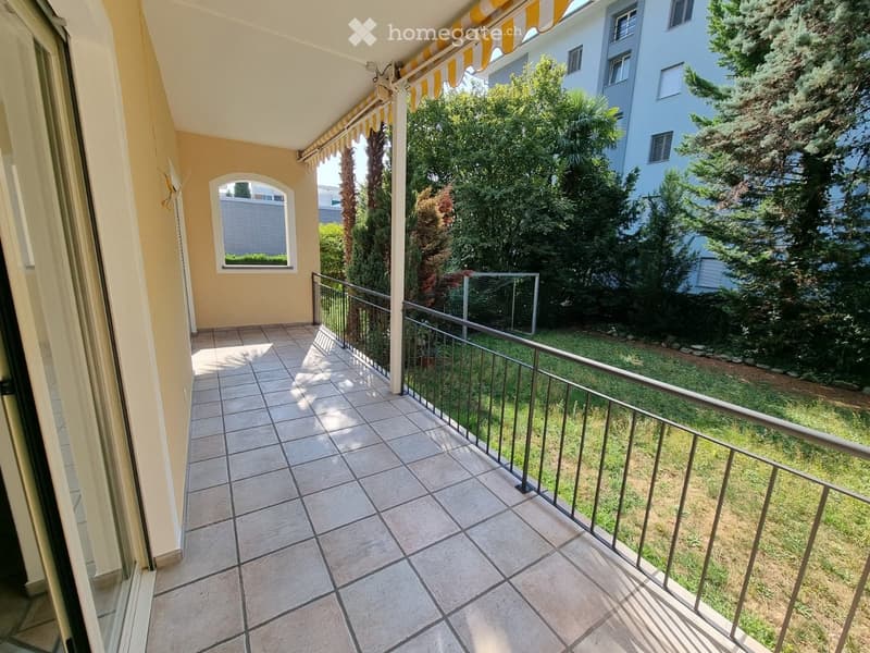 Balcony with tiled floor, metal railing, and striped awning, overlooking a green garden area with trees and a neighboring building.