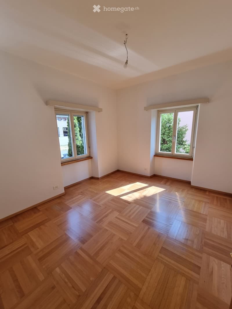 Empty room with wooden flooring, white walls, and large windows with wooden sills. Natural light streams in, illuminating the space.