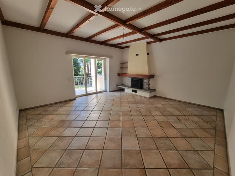 Empty room with tiled floor, exposed wooden ceiling beams, fireplace with stone surround, and sliding glass door leading to a balcony.
