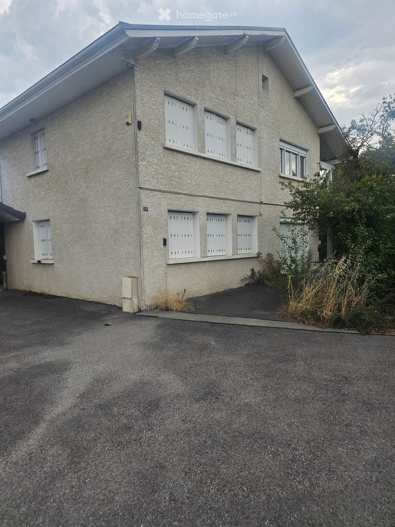 Two-story house with light gray textured walls, white-framed windows, and a flat roof with white trim. The house has a small garden area with shrubs and a paved driveway.