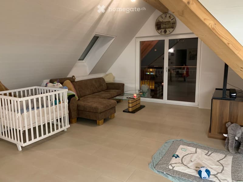 Bright attic living space with slanted wooden ceilings, large windows, and a sliding glass door leading to a balcony. Features a brown sectional sofa, glass coffee table, baby crib, and decorative toys.