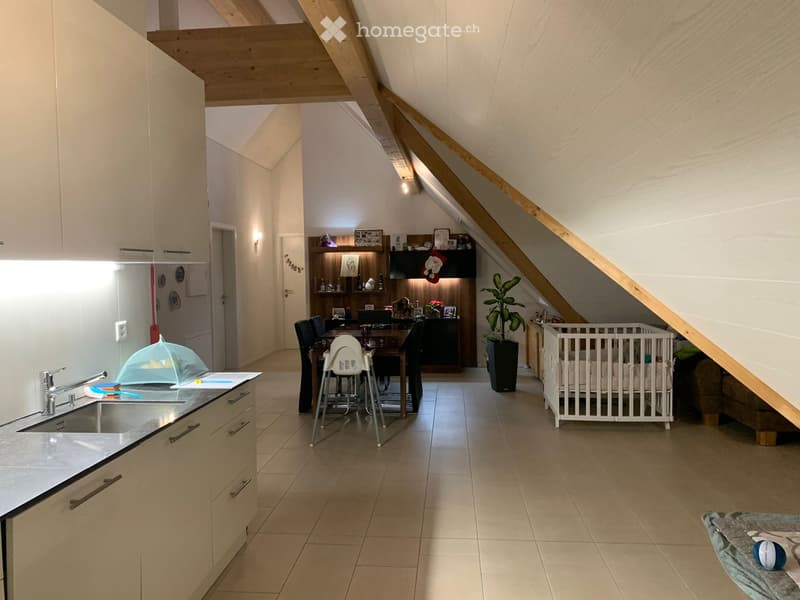 Open-plan attic space with kitchen area, dining table, baby crib, and living area. Features exposed wooden beams, modern cabinets, and a potted plant.