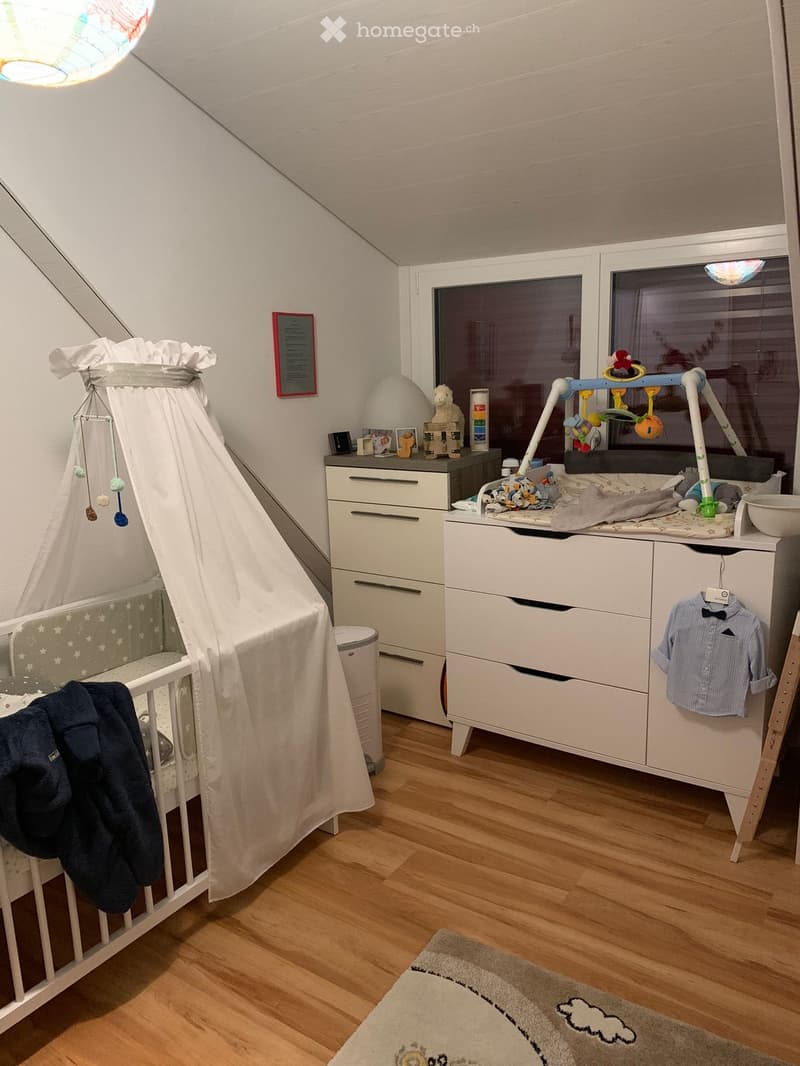 A well-lit children's bedroom with a crib, changing table, drawer unit, and various toys. The room features wooden flooring, a rug, and a slanted ceiling typical of an attic space.