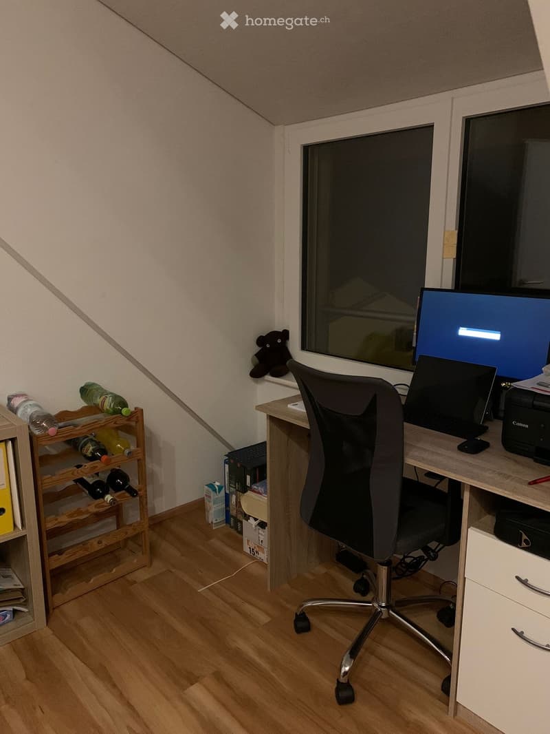 A small, well-lit room with wooden flooring, a desk with a laptop, chair, and printer, a bottle rack, and a window with a view.