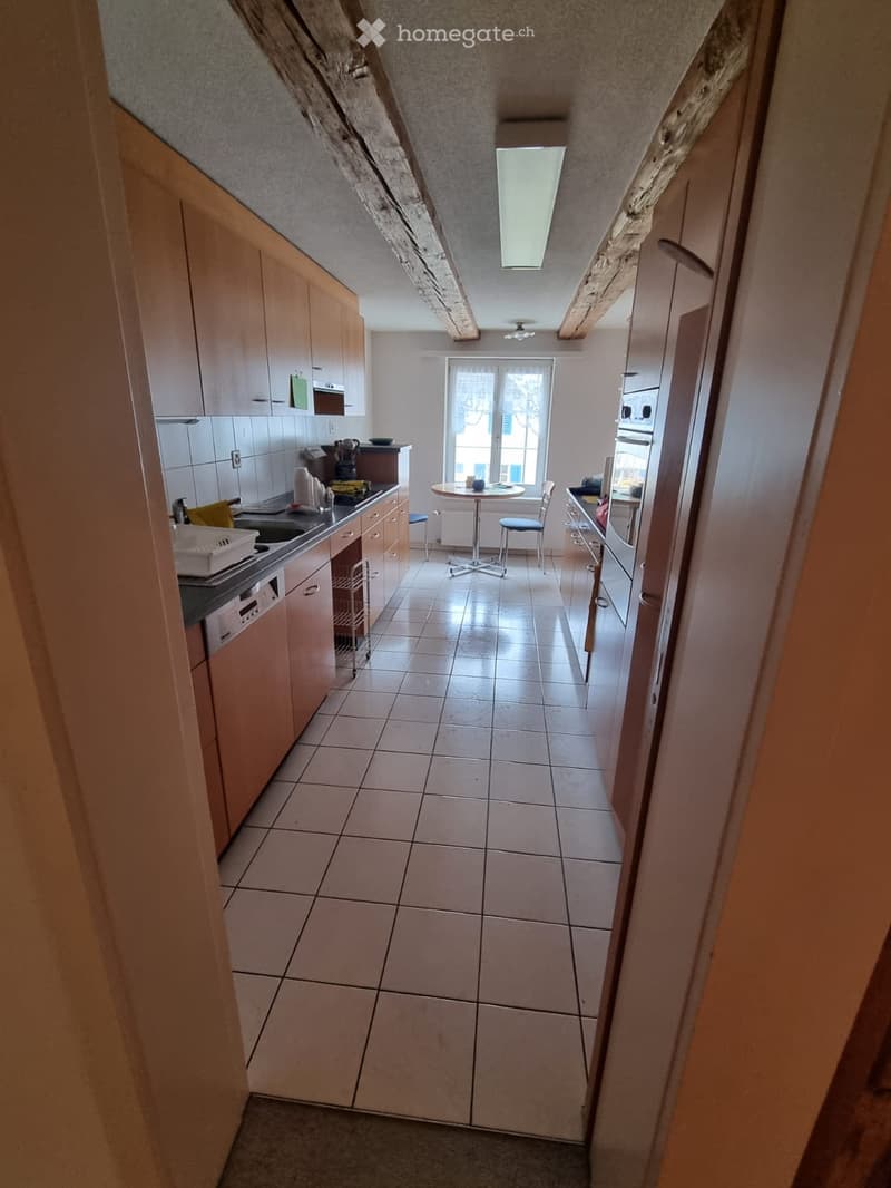 Modern kitchen with light wooden cabinets, tiled floor, and stainless steel appliances. Features a small round table with two chairs near a window.