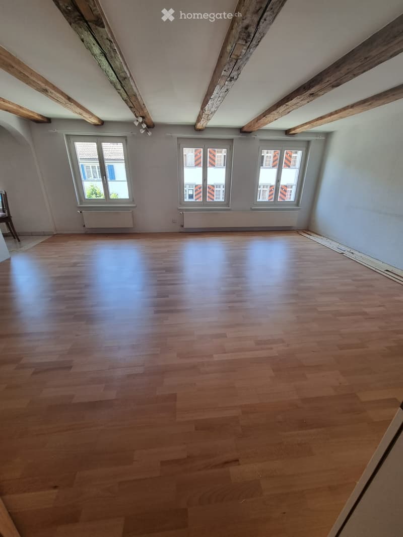 Empty room with wooden flooring, exposed wooden beams on the ceiling, and three windows with white frames and grey blinds.
