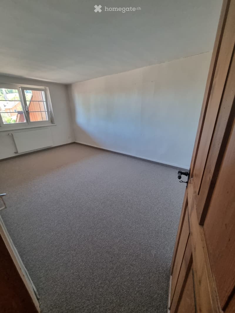 Empty room with light grey walls, carpeted floor, and a single window with white frame and metal bars. No furniture or equipment visible.