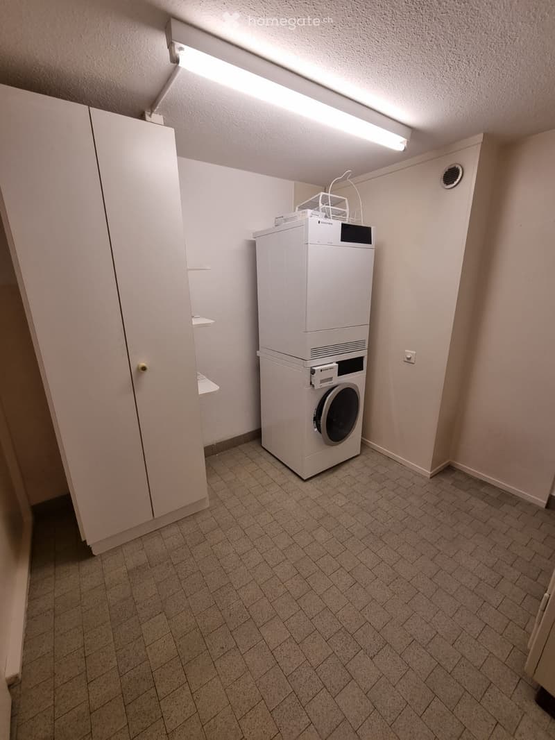 A small, well-lit room with tiled flooring and a white washing machine positioned against the wall.