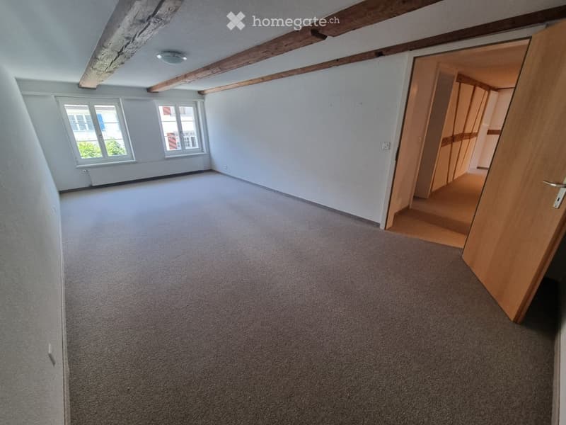 Empty room with grey carpet, white walls, wooden beams on the ceiling, and two windows letting in natural light. A wooden door leads to another room.