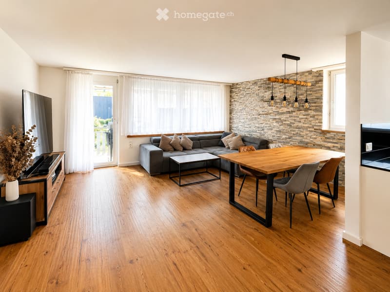 Modern living and dining area with wooden flooring, grey sectional sofa, wooden dining table with chairs, TV, and decorative vase with dried flowers.