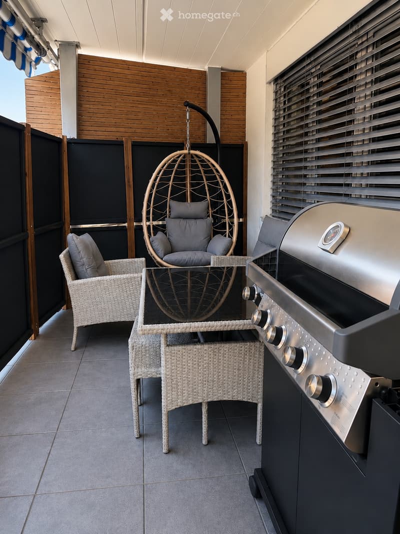 Balcony with a wicker hanging chair, a wicker chair, a small table, and a grill. The space features wooden paneling and a covered ceiling.