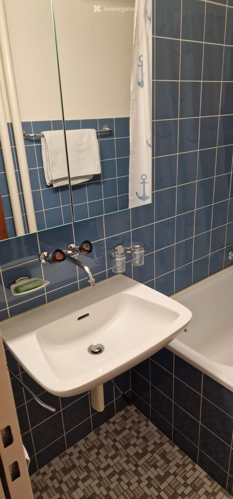 Bathroom with a white sink, chrome faucet, mirror, towel, and a bathtub. Blue tiles on walls and a patterned tile floor.