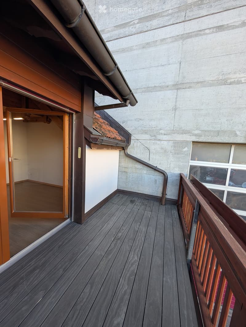 Wooden balcony with dark grey decking, wooden railing, and a view of a concrete wall and adjacent room.