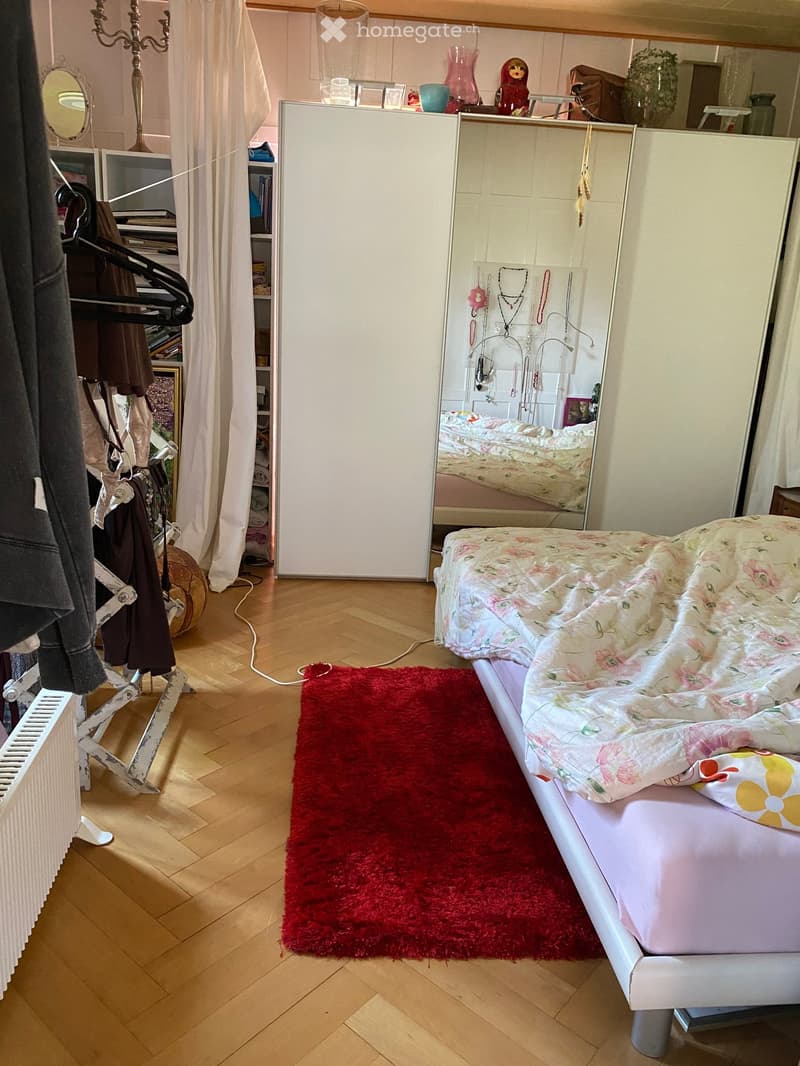 A well-lit bedroom with a wooden floor, a white bed with floral bedding, a red shag rug, a large mirror, a wardrobe, and a clothing rack with hanging clothes.